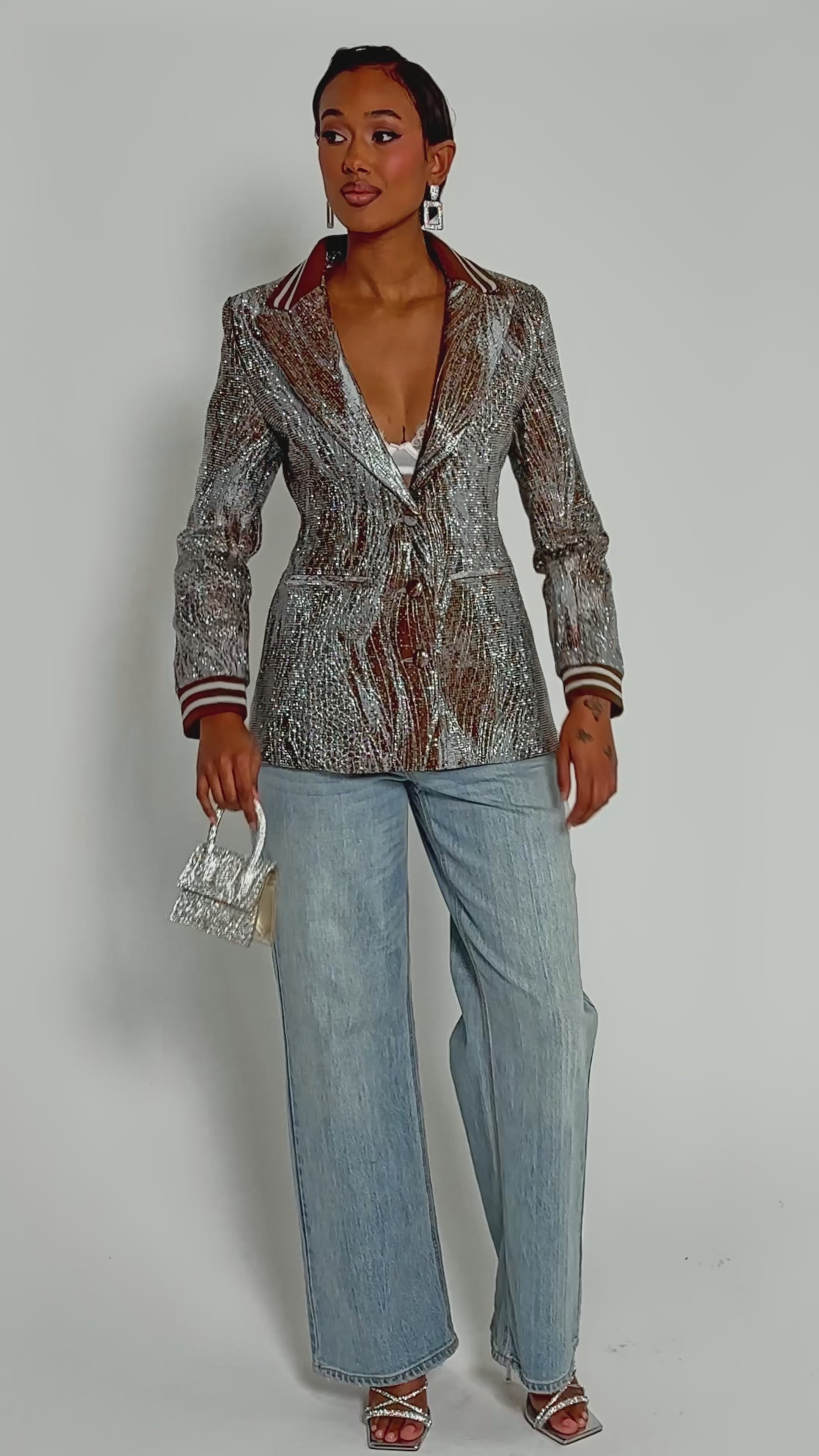 Video of woman modeling the DASH EMBELLISHED BROCADE BLAZER, a shiny silver and brown metallic piece, styled over a white lace top with light blue jeans, geometric earrings, and rings.