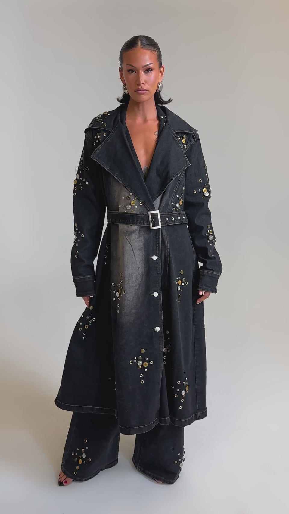 A video of a woman in the SPACE AND TIME STUDDED DENIM TRENCH and matching wide-leg pants, both detailed with metallic grommets, against a simple backdrop.