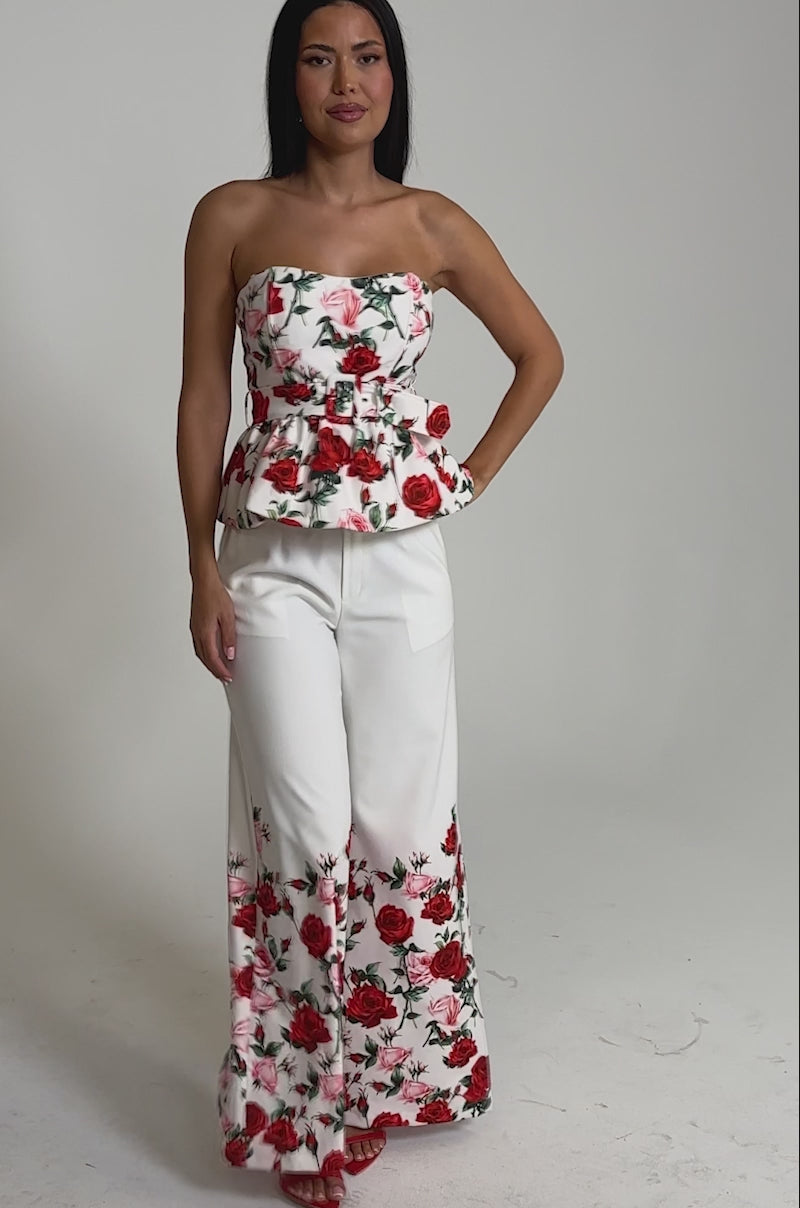 A video of a woman in the FULL BLOOM FLORAL TROUSER, featuring white high-rise wide-leg pants with red rose prints, paired with a matching strapless peplum top and red high-heeled sandals.