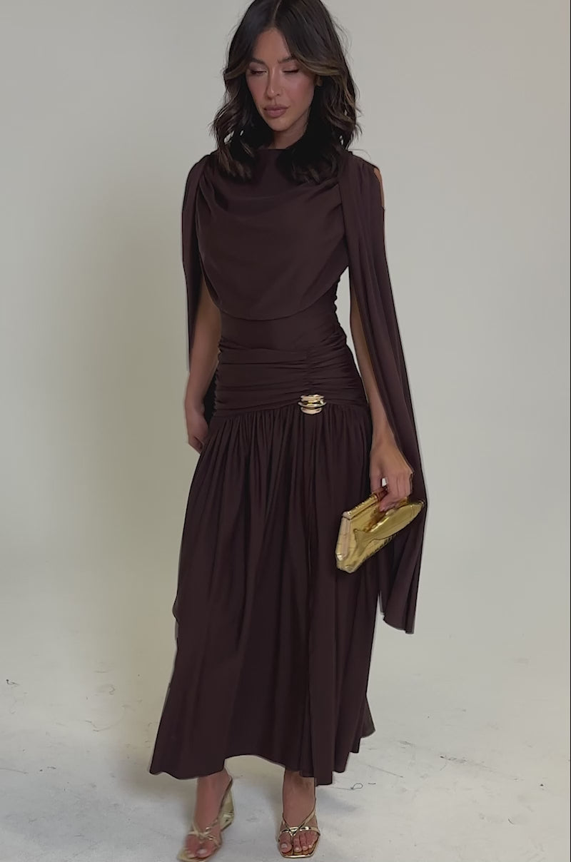 Video of woman modeling the HILLARY SEAMLESS BODYSUIT in chocolate brown, featuring a draped design with flowing sleeves and a high neckline. She accessorizes with a gold clutch and heels, set against a simple light background.