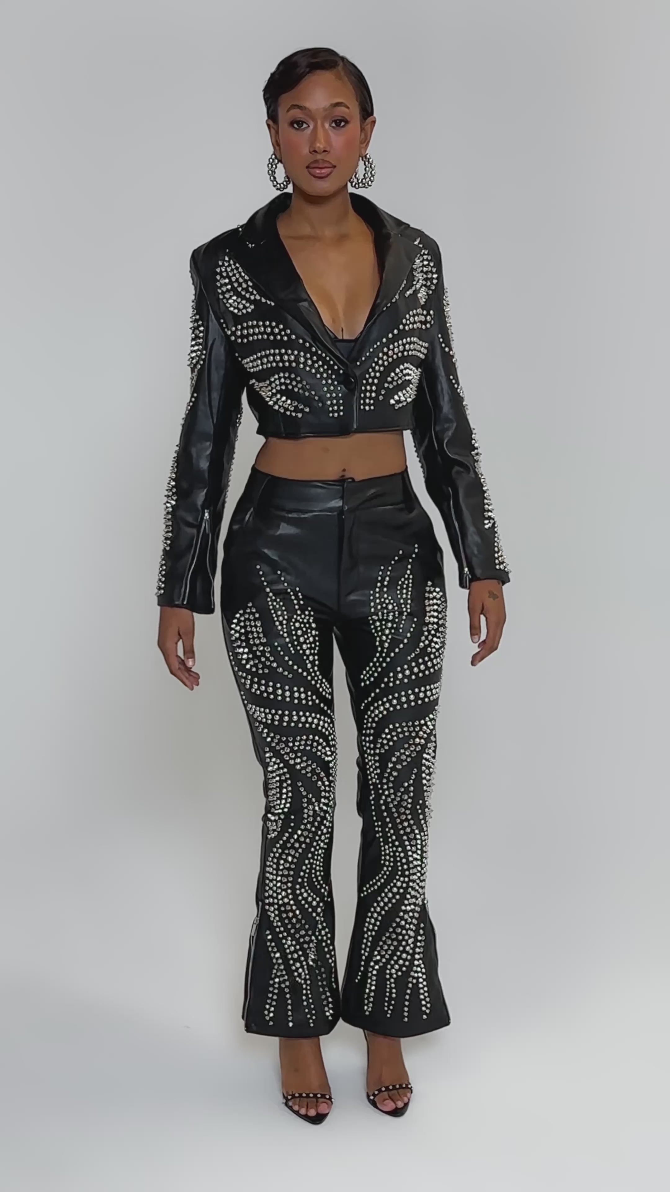Video of woman modeling the GONE BAD STUDDED PANT, a pair of black high-rise flare pants with swirling stud patterns, styled with a matching studded crop jacket, black open-toe heels, and hoop earrings.