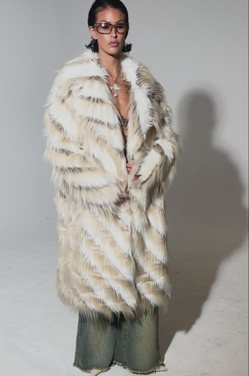 A video of a model wearing sunglasses and the HARLOW PATCHWORK FAUX FUR coat with black and white stripes, loose jeans, and displaying chest tattoos, someone poses confidently against a plain background.