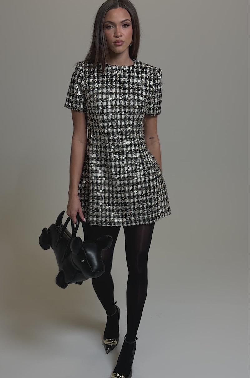 Video of woman modeling the GIRLY POP TWEED MINI DRESS, featuring a black and white checkered pattern and subtle sequin details. She stands against a plain background, holding a black handbag.