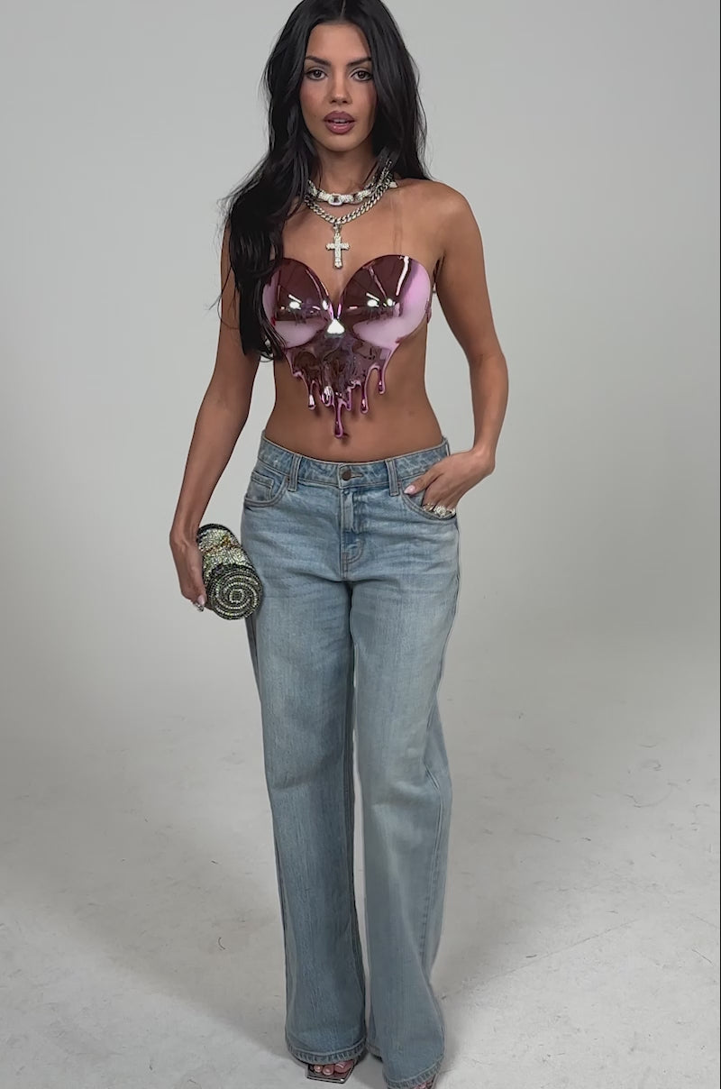 Video of woman modeling the DRIPPED OUT TOP, a strapless pink metallic heart-shaped piece with a dripping effect, layered necklaces, light blue jeans, and holding a rhinestone clutch for a striking nightlife look.