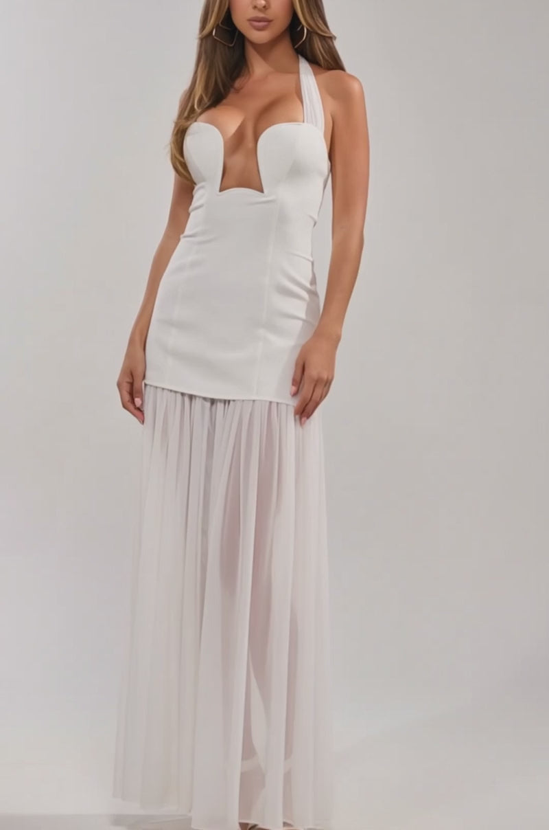 Video of woman modeling the ANGEL ENERGY MAXI DRESS IN WHITE featuring a halter neck, deep neckline, fitted bodice, and sheer flowing skirt. She has long wavy hair, layered necklaces, and stands against a plain light background.