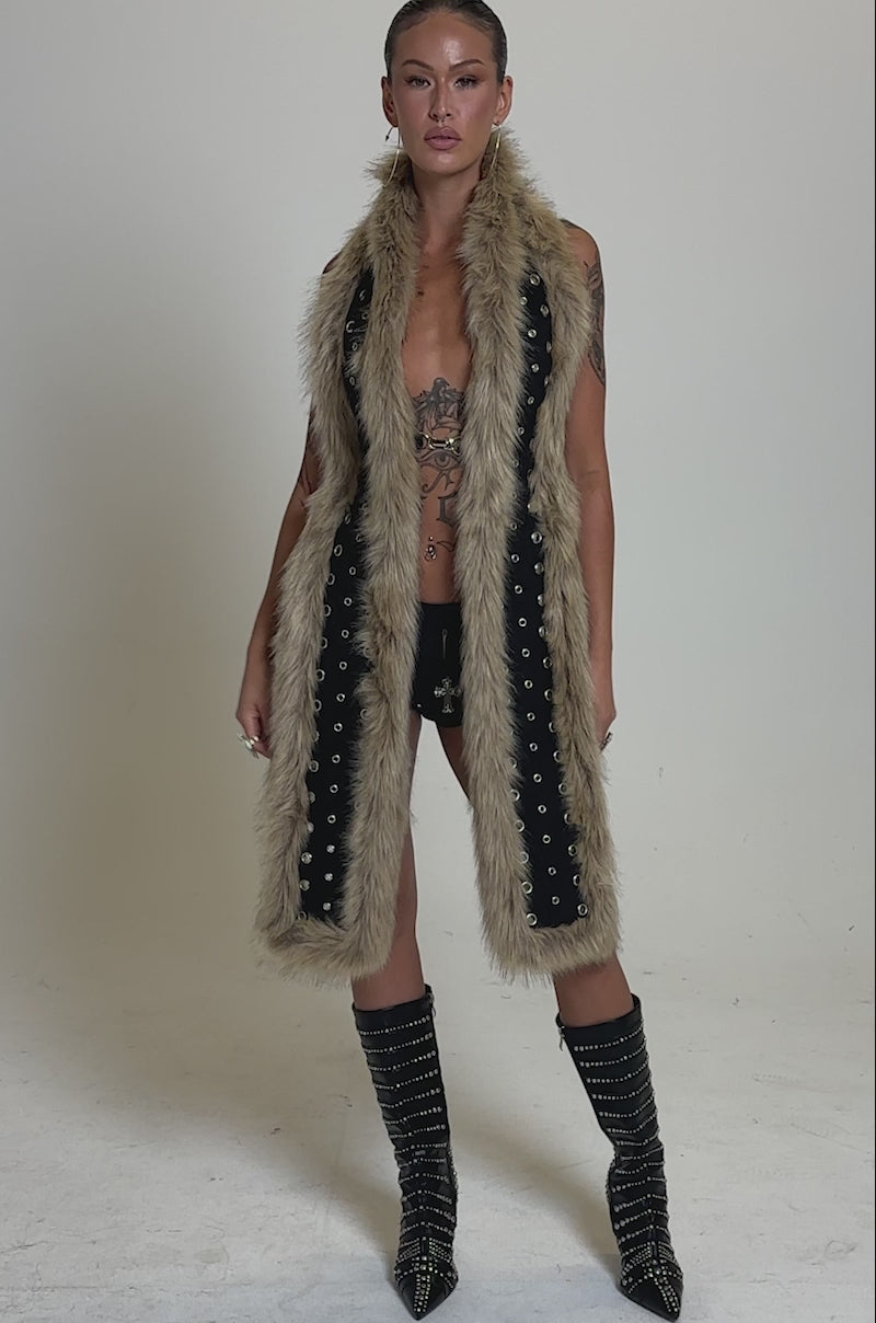 A video of a person standing confidently against a neutral gray background, wearing the ICY HOT FUR BRALETTE, black shorts, and knee-high black boots with silver studs. 