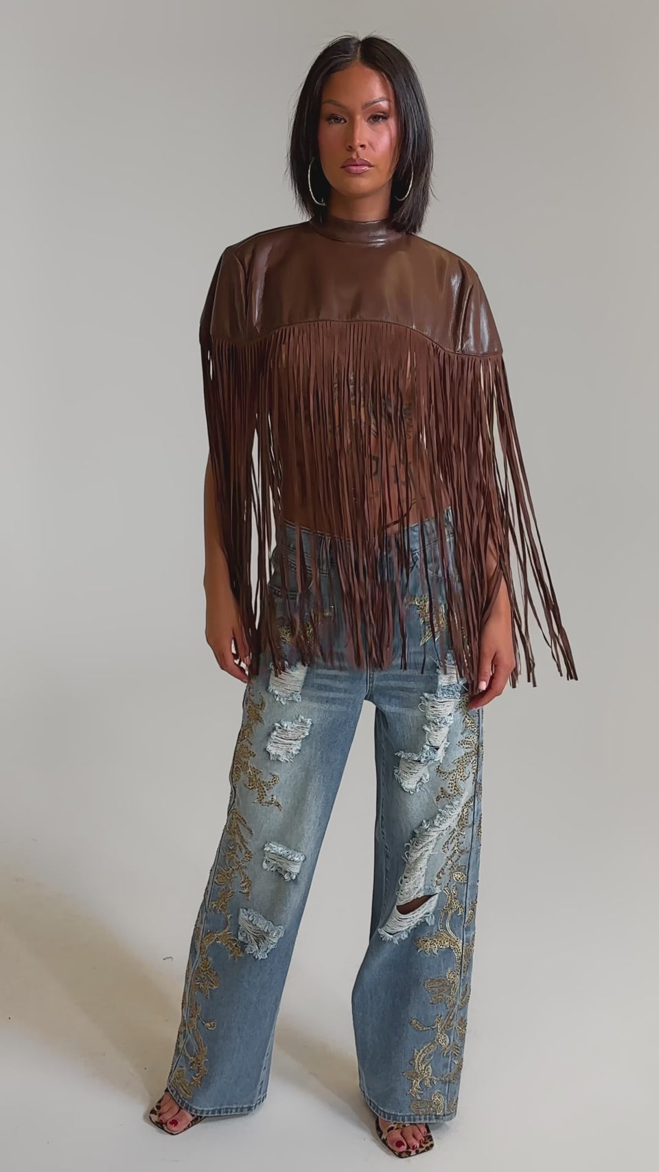 A video of a woman wearing the FRINGE FOR DAYS FAUX LEATHER TOP, a brown festival layering piece with fringe, styled with wide-leg ripped jeans featuring gold embroidery against a plain light background.