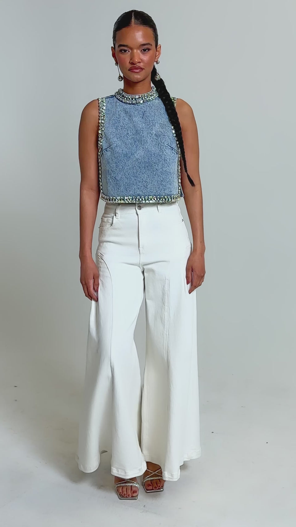 Video of woman modeling the POPPING OUT RHINESTONE DENIM BLOUSE, a sleeveless, high-neck crop top with silver embellishments. She pairs it with high-waisted white pants and dangling earrings, her slicked-back hair enhancing the blouse’s modern edge.