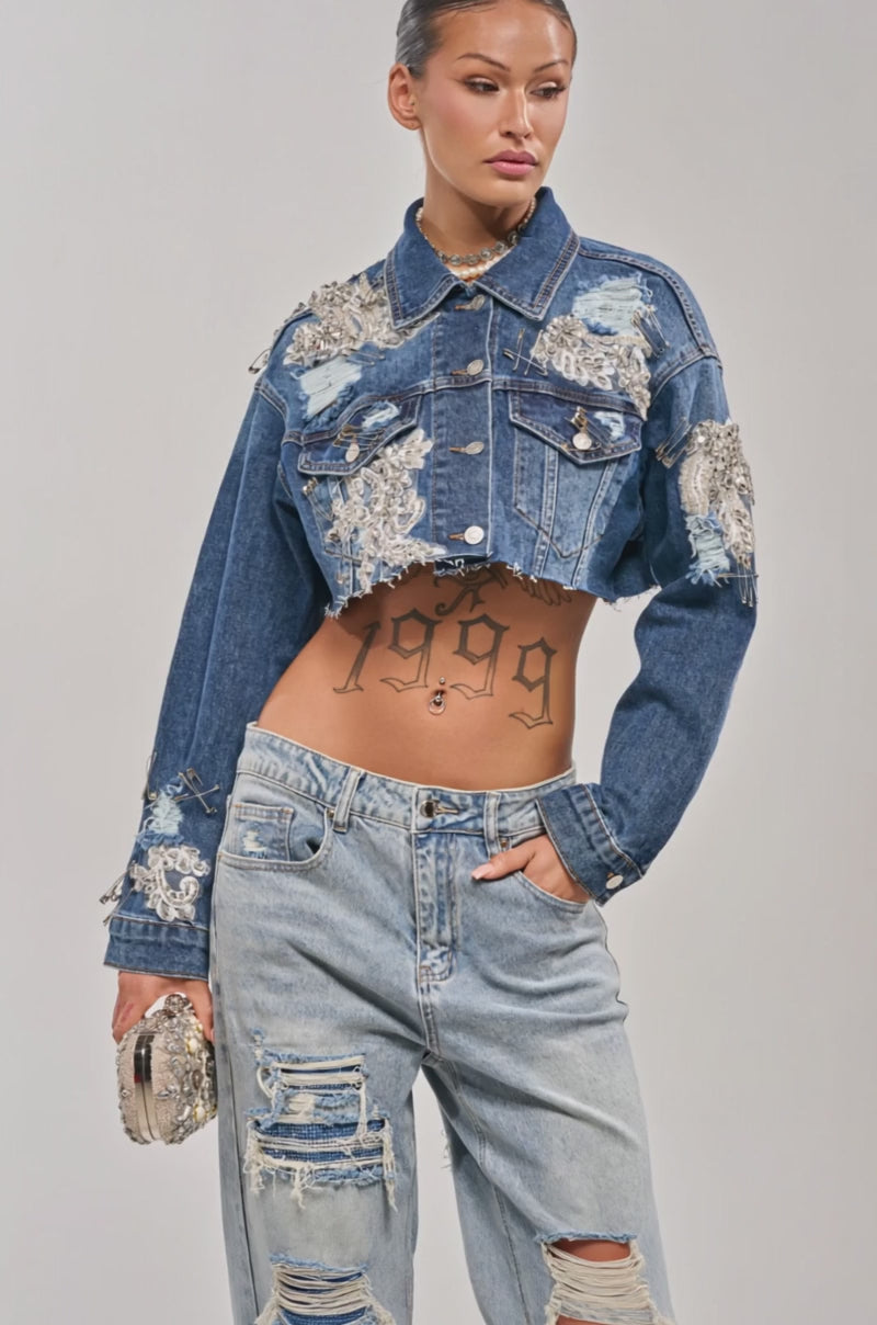 Video of woman modeling the LET'S RIDE EMBELLISHED DENIM JACKET, a cropped rhinestone embroidered denim piece over a white bralette with matching jeans. Layered necklaces and bold tattoos accent her chest and stomach.