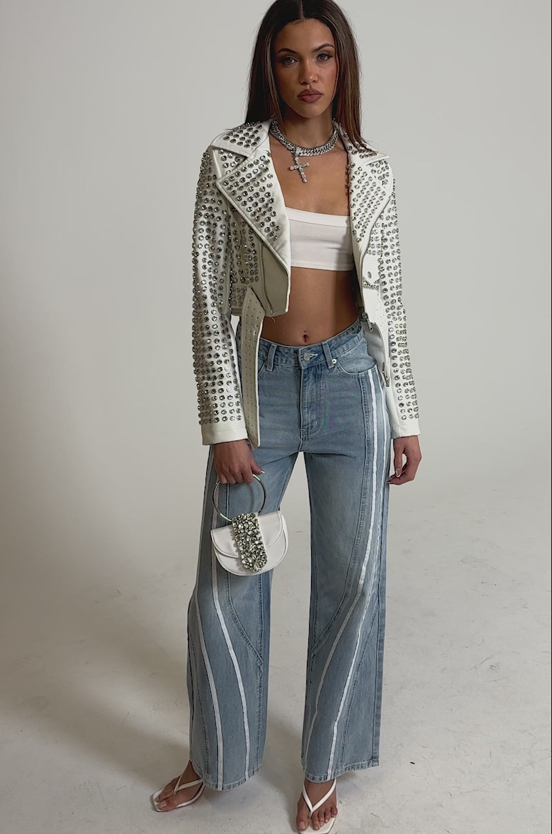 Video of woman modeling the SHE IS A RUNNER DENIM TRACK PANTS with double stripe side panels, a white studded cropped jacket, white heeled sandals, layered necklaces, and holding a small white purse against a plain light gray background.