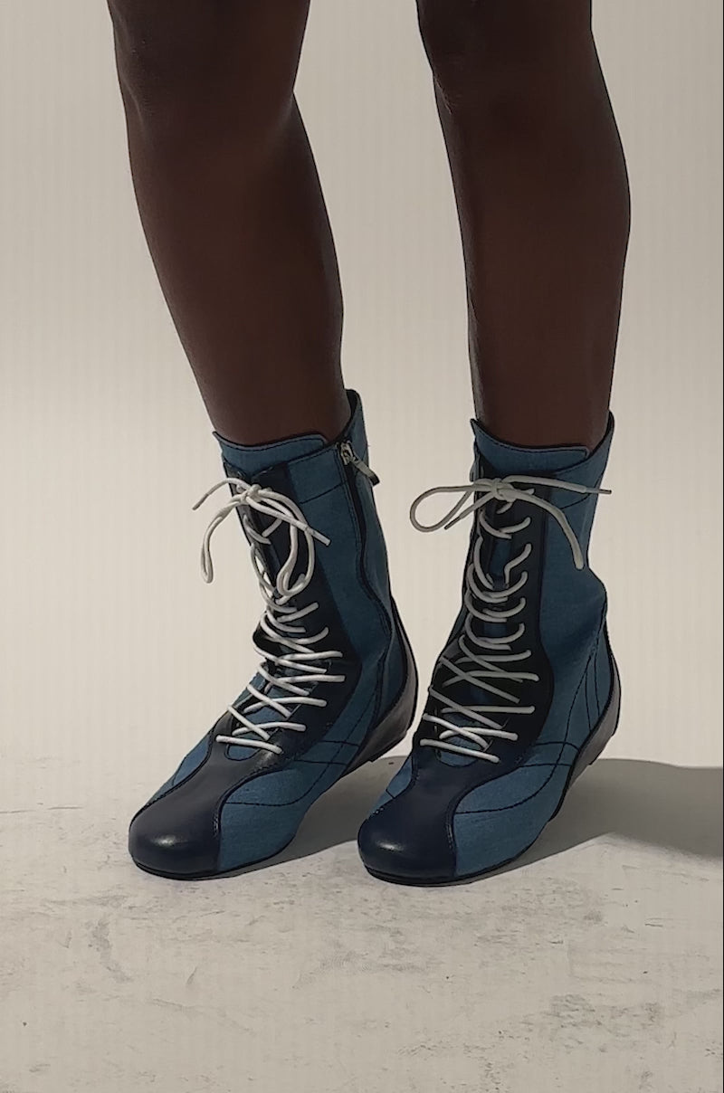 A video of a model wearing the AZALEA WANG PASSER NAVY FLAT SNEAKER: knee-high, denim blue lace-up boots with white laces and side zippers in a sneaker style. 