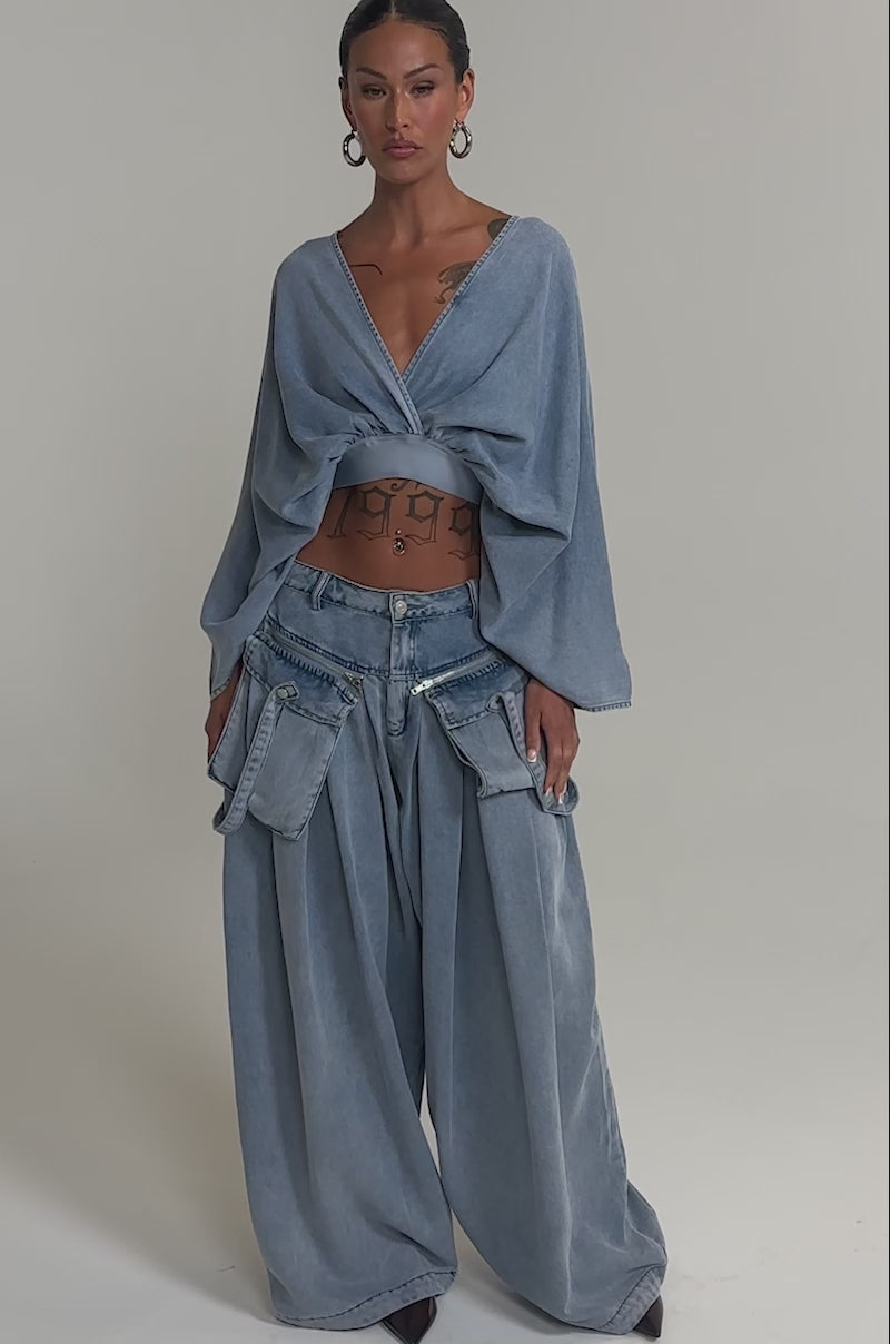 Video of woman modeling the DRIFTING AWAY BLOUSE IN LIGHT BLUE DENIM with baggy blue jeans, large hoop earrings, and creates an elegant day-to-night look.
