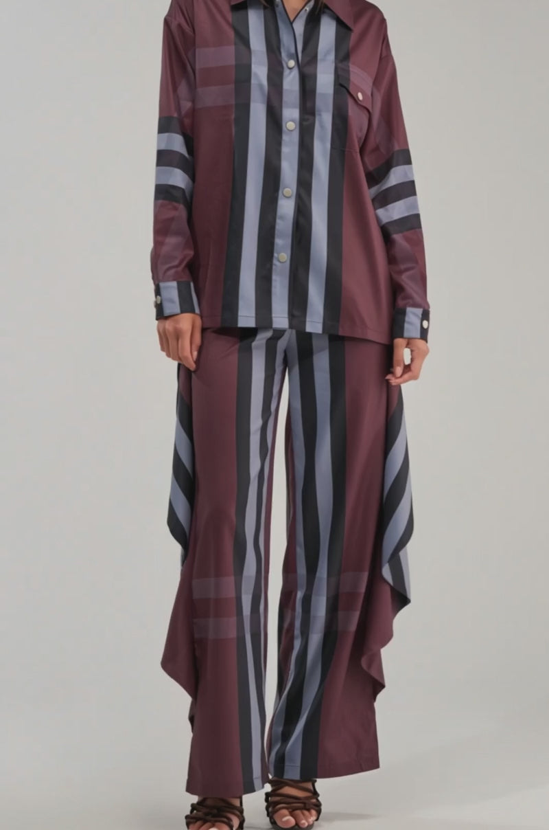 A video of a woman in a relaxed silhouette, wearing the LUXURY LIFE PLAID BUTTON DOWN with loose, long sleeves, paired with wide-leg striped pants featuring draped side panels and black heeled sandals.