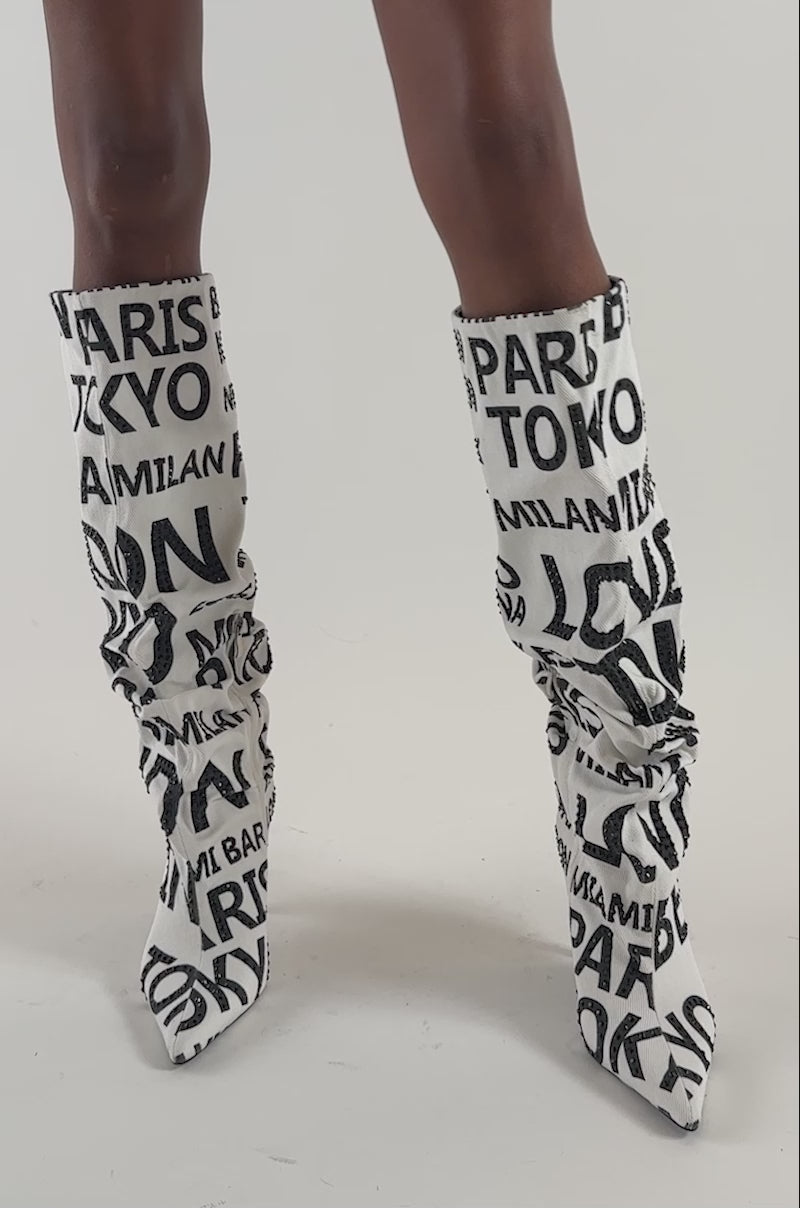 Video of a woman modeling the AKIRA GOLD GLENN BLACK WHITE CITIES STILETTO BOOT—a striking white knee-high boot adorned with bold black city names like London, Paris, and Tokyo. 