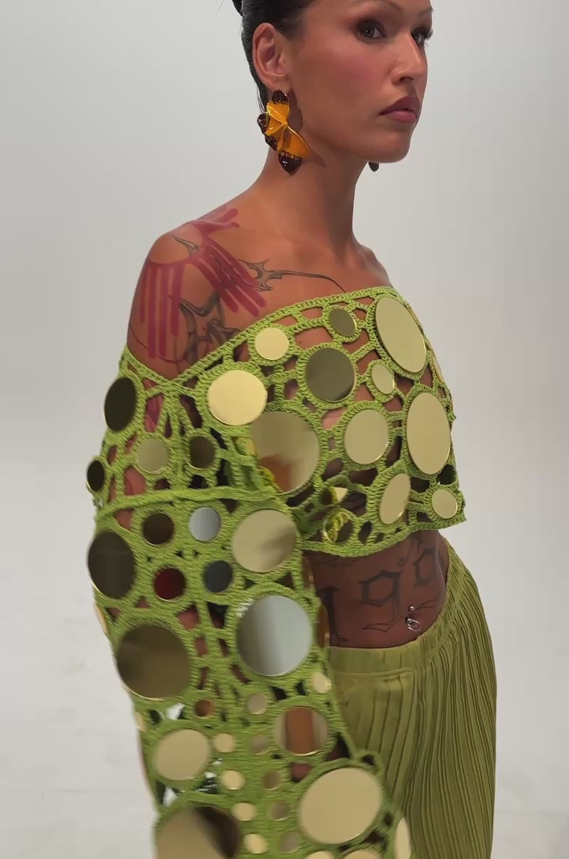 Video of a woman modeling the RESORT VIBES CROCHET CROP LONG SLEEVE IN CHARTREUSE paired with green pleated pants. The vibrant off-the-shoulder top features bold gold and green circular accents, complemented by striking earrings and visible tattoos—an ideal vacation-ready look.