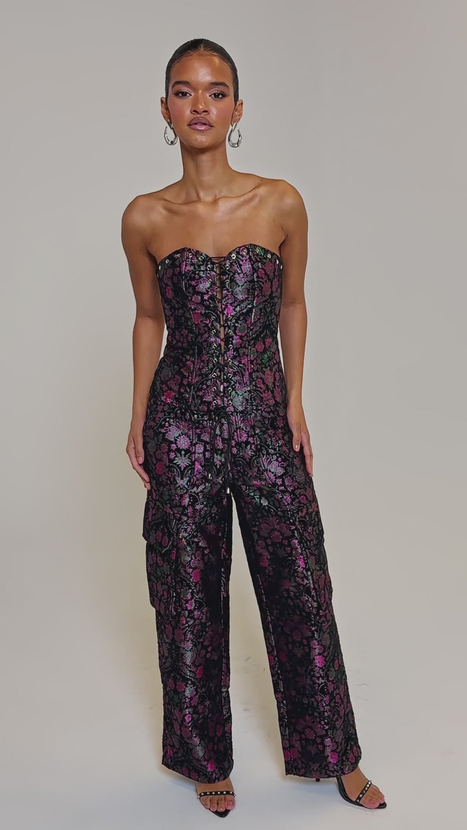 Video of woman modeling the KIMMY FLORAL TROUSER with pink and purple floral print, high-rise flared legs, and pocket details. She pairs it with slicked-back hair, hoop earrings, and open-toe heels.