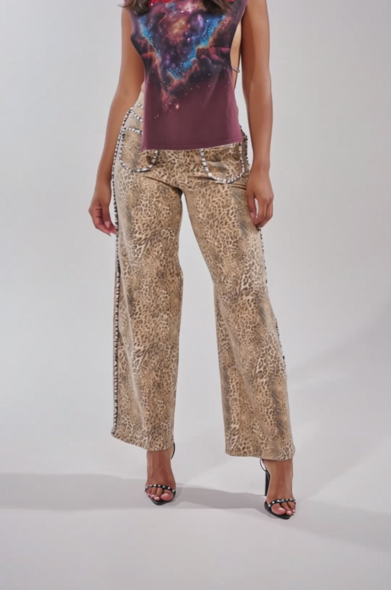A video of a woman in IN THE WILD LEOPARD DENIM wide-leg pants, a sleeveless graphic top with zebra-print trim, and black heeled sandals.