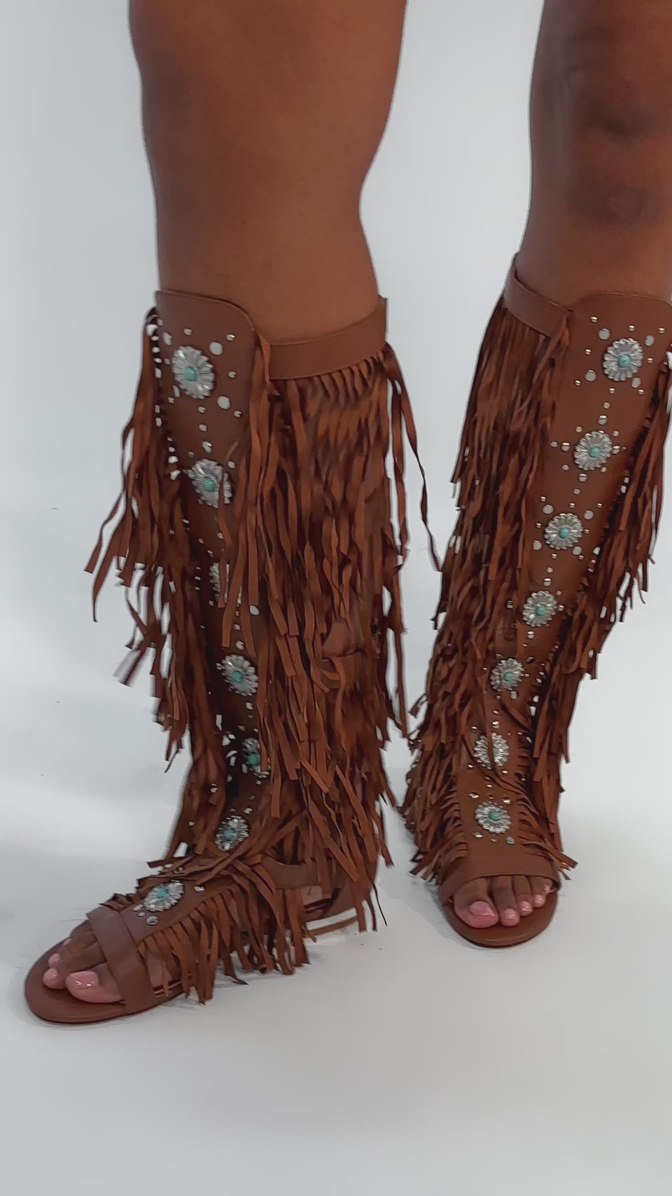 Video of woman modeling the AZALEA WANG DUSTINE BROWN GLADIATOR EMBELLISHED SANDAL, featuring knee-high straps with turquoise and silver details, while standing on a light background.