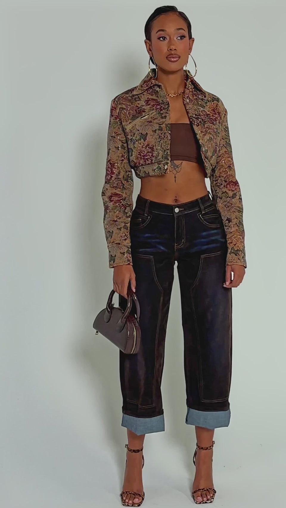 Video of woman modeling the NOW OR NEVER TAPESTRY MOTO jacket with high-waisted dark jeans poses confidently with one hand on her hip against a plain background.