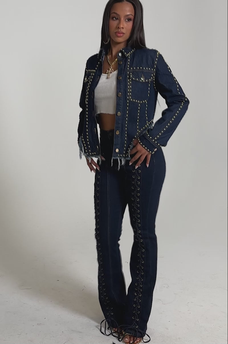 A video of a woman in the HOTLANTA STUDDED DENIM JACKET and matching high-waisted jeans with grommet and lace-up accents, paired with a white crop top and strappy heels. 