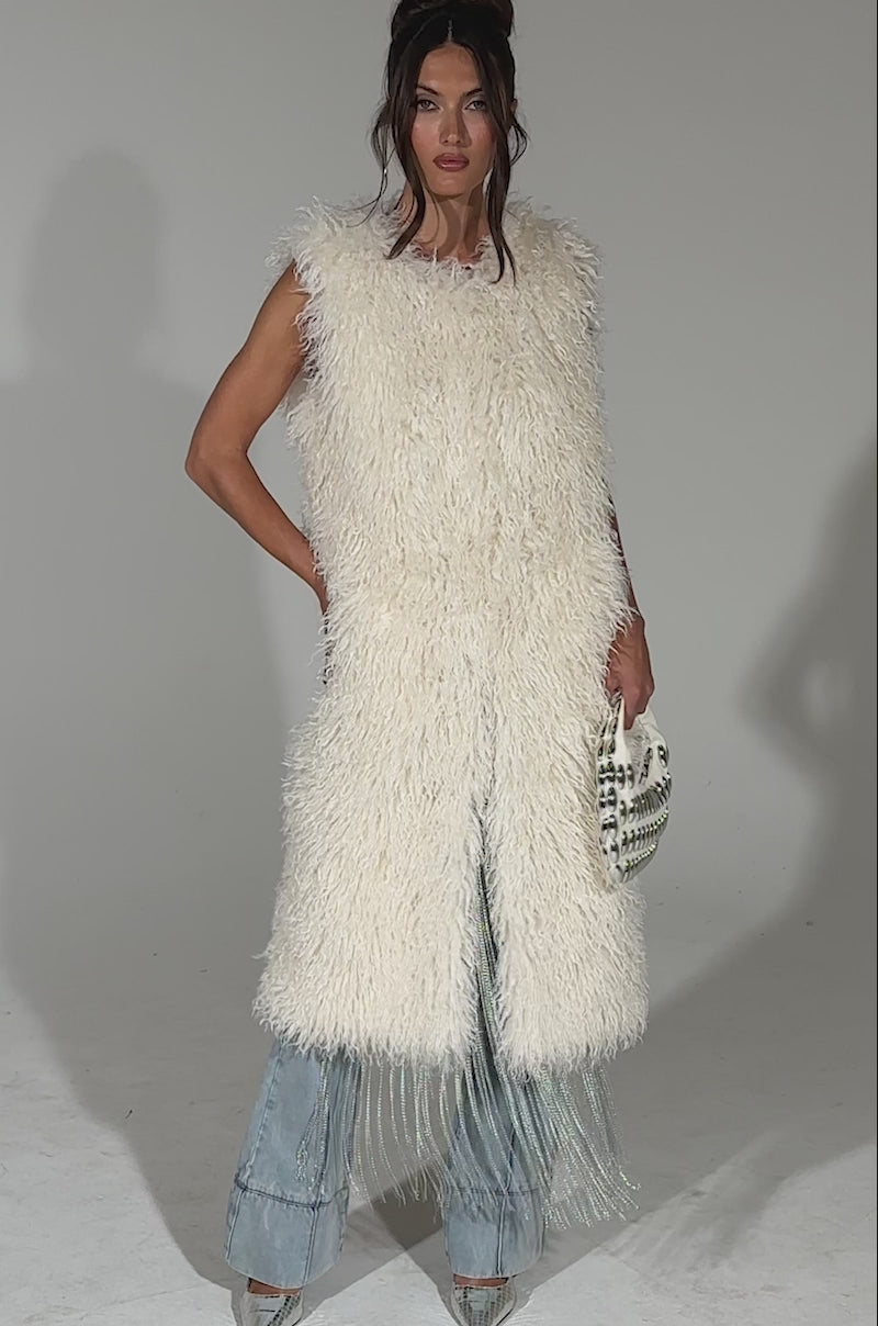 A video of a woman with long brown hair in an updo wearing the sleeveless white ANOTHER LANE MOHAIR VEST and holds a small white handbag, standing against a plain light blue background.
