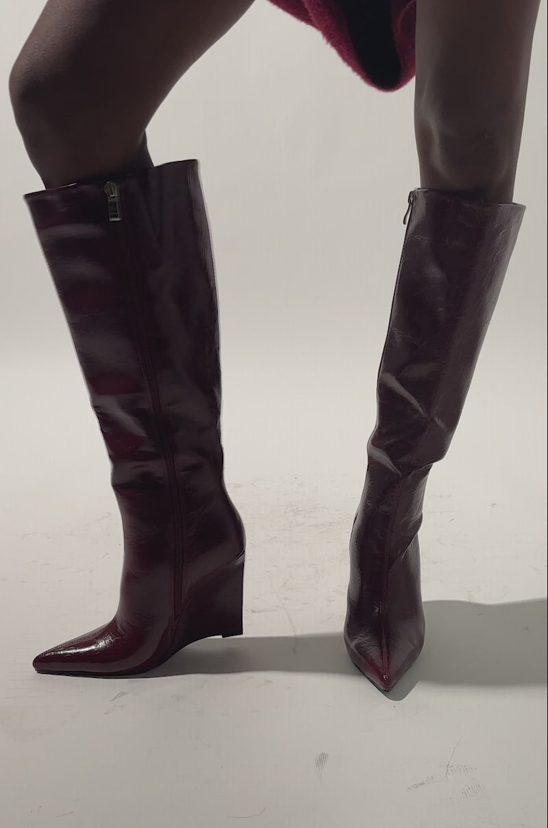 A video of a person wearing the AZALEA WANG AMAADIS BURGUNDY WEDGE KNEE HIGH BOOT, featuring shiny wedge heels, and a textured red garment, stands on a light background.