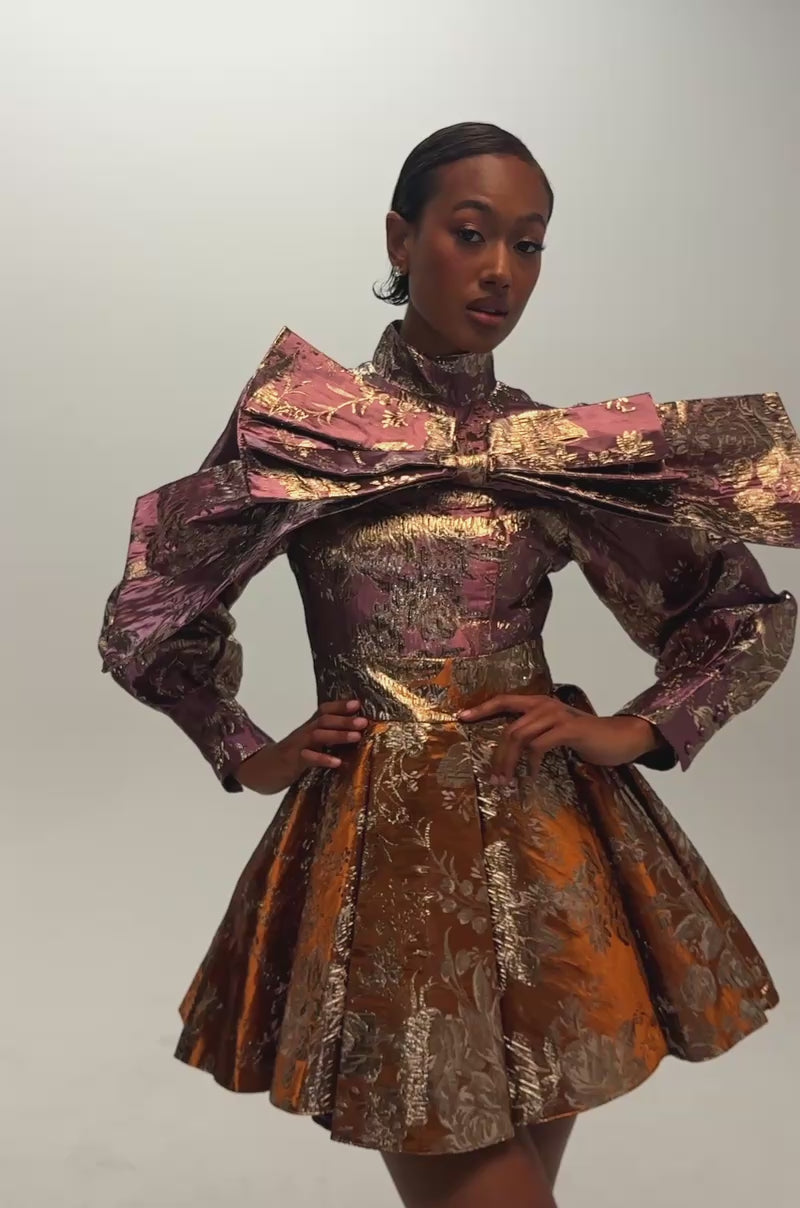 Video of a woman modeling the RENAISSANCE TWO TONED BROCADE MINI DRESS IN PURPLE COMBO—featuring a metallic, floral-patterned brocade fabric, exaggerated puff sleeves, a large bow detail on the chest, and a flared skirt in shades of purple and bronze.