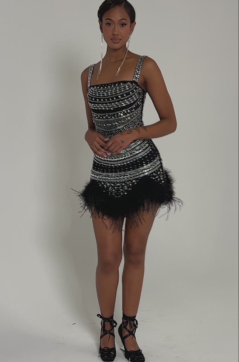 Video of woman modeling the SHOWSTOPPER RHINESTONE MINI DRESS, featuring a sparkling geometric pattern, feather-trimmed hem, and long dangling earrings, set against a softly lit gray background.