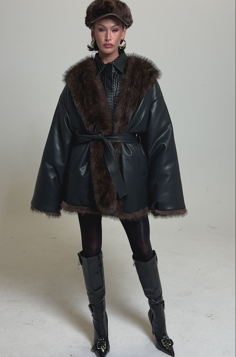 A video of a person in the CAN I KICK IT REVERSIBLE PUFFER, styled with a brown fur hat, tall textured boots, and dark tights against a plain background.