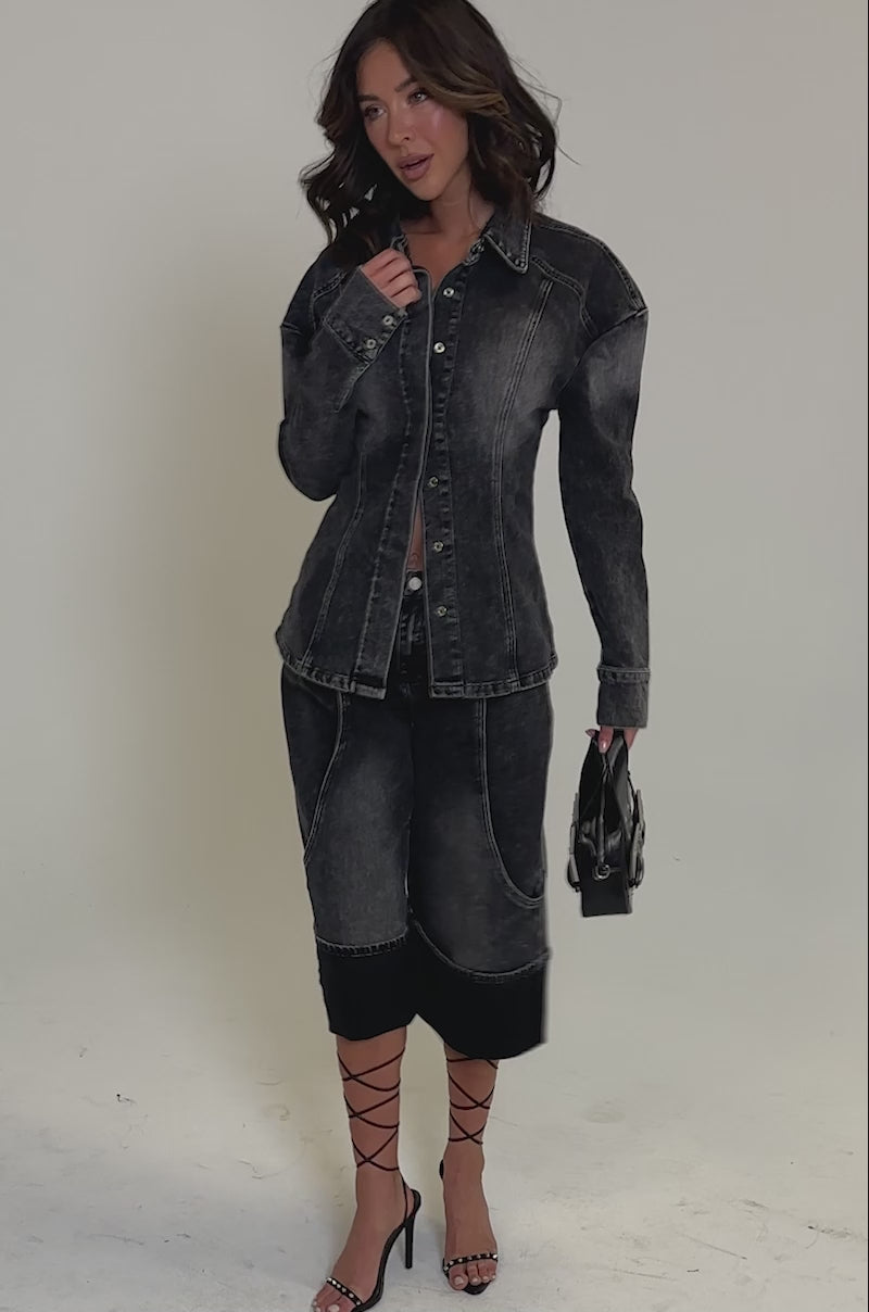 Video of woman modeling the IN MY OWN WORLD BUTTON DOWN, a black acid-wash denim jacket with paneled design and matching pants, against a plain backdrop, holding a small black handbag 