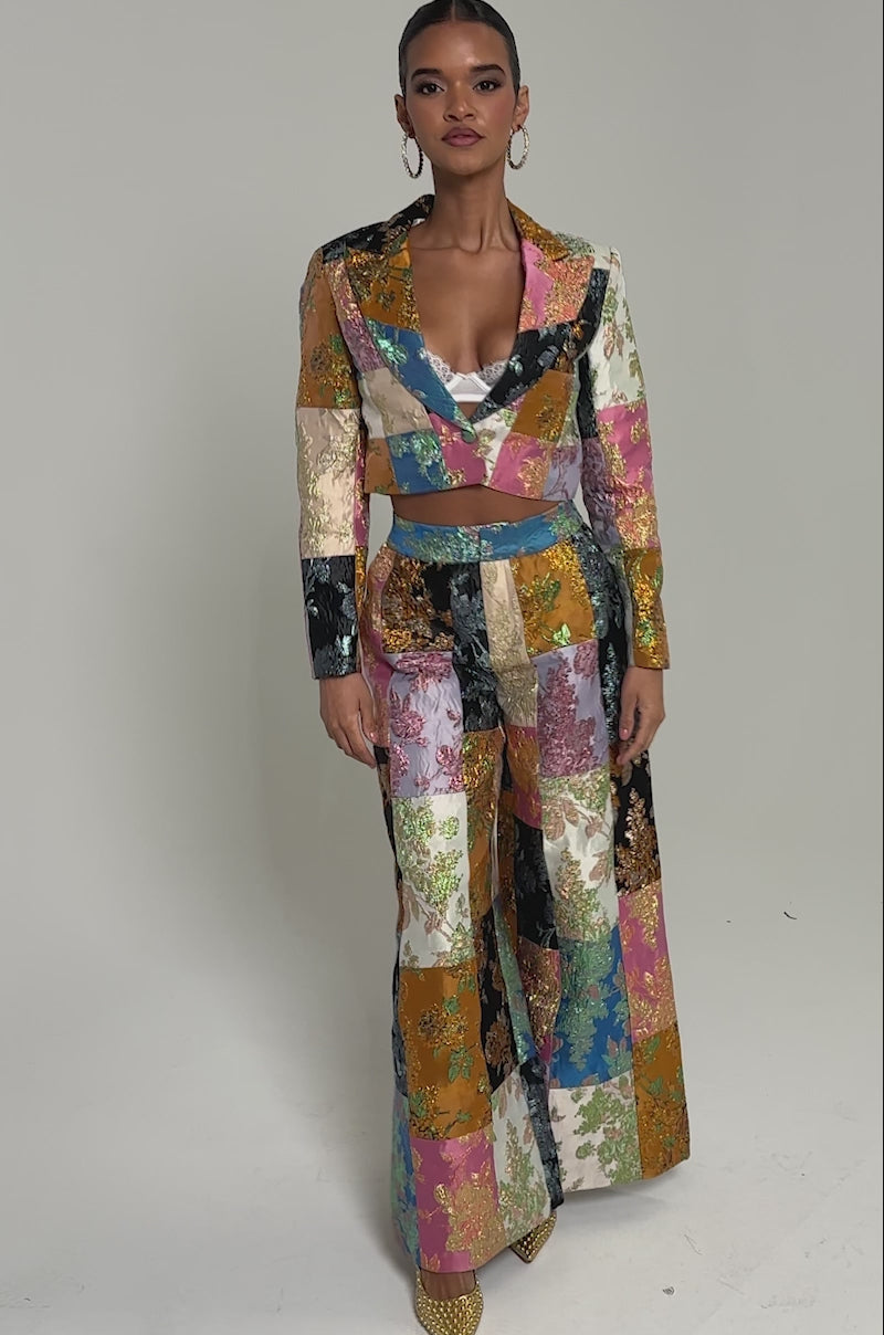 Video of woman modeling the LOLITA PATCHWORK BROCADE BLAZER with intricate floral patterns and matching pants. Her slicked-back hair and bold hoop earrings complete the look, set against a plain background.