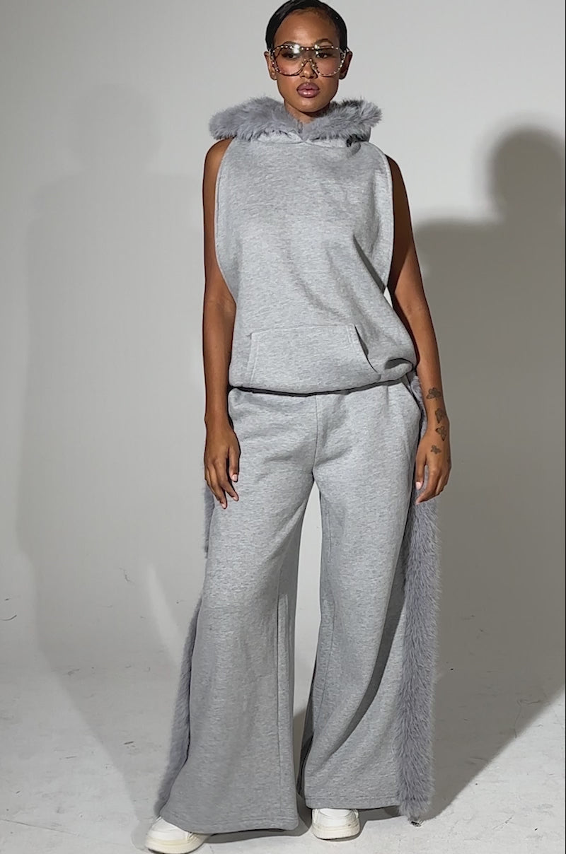 A video of a model wearing a sleeveless grey hoodie, sunglasses, white shoes, and the LUXE FUR JOGGER IN GREY with fur side trim, a person stands against a plain light background.