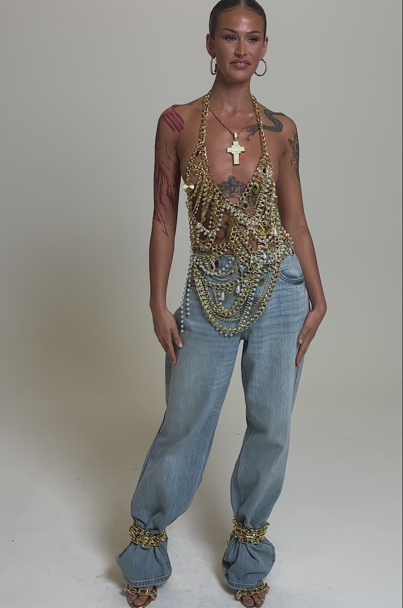 Video of woman modeling the DRIP DRIP BRALETTE—gold chain style layered with pearl necklaces—as a standout festival top, paired with light blue jeans, hoop earrings, and styled hair against a gray background. Tattoos are visible.