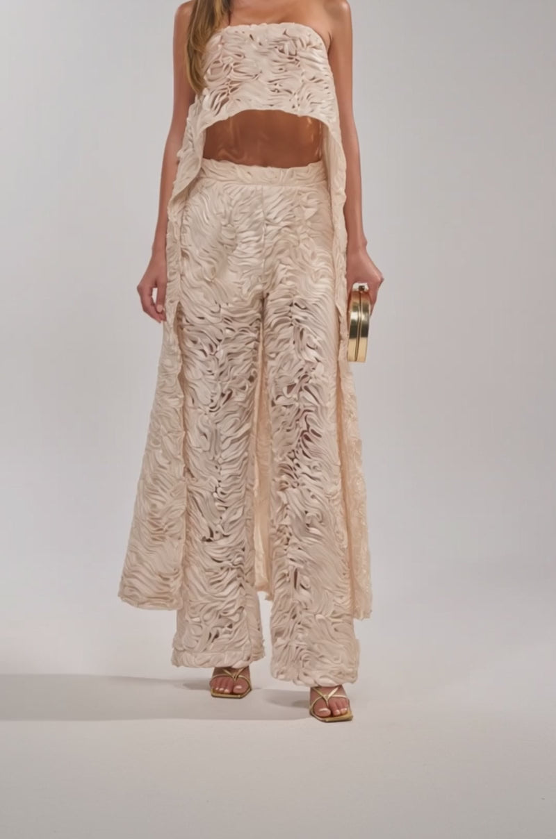 A video of a woman wearing the MAEVE SLEEVELESS BLOUSE paired with textured cream wide-leg pants, a gold clutch, and gold high-heeled sandals.