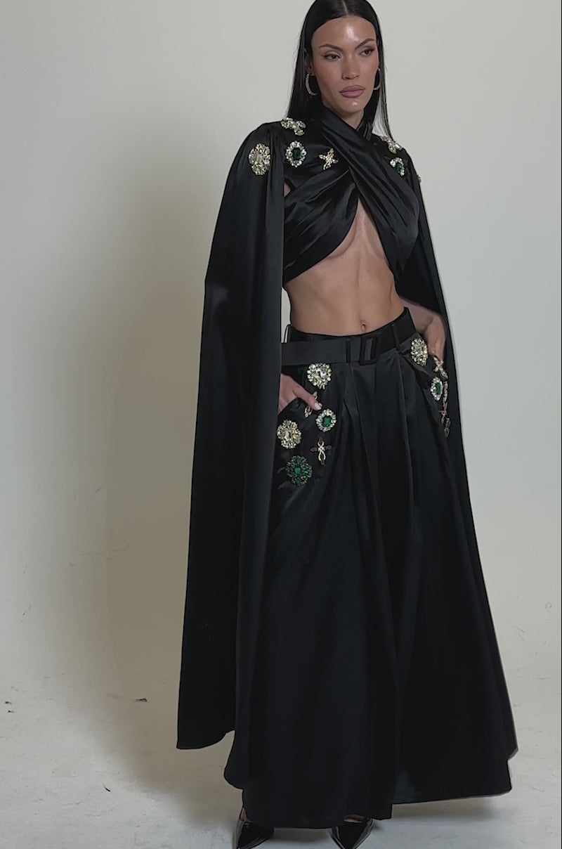 A video of a woman in the AFTER DARK BLOUSE and wide-legged pants with colorful embroidered patches, accessorized with hoop earrings and black heels. She has long straight hair and stands against a plain light background.