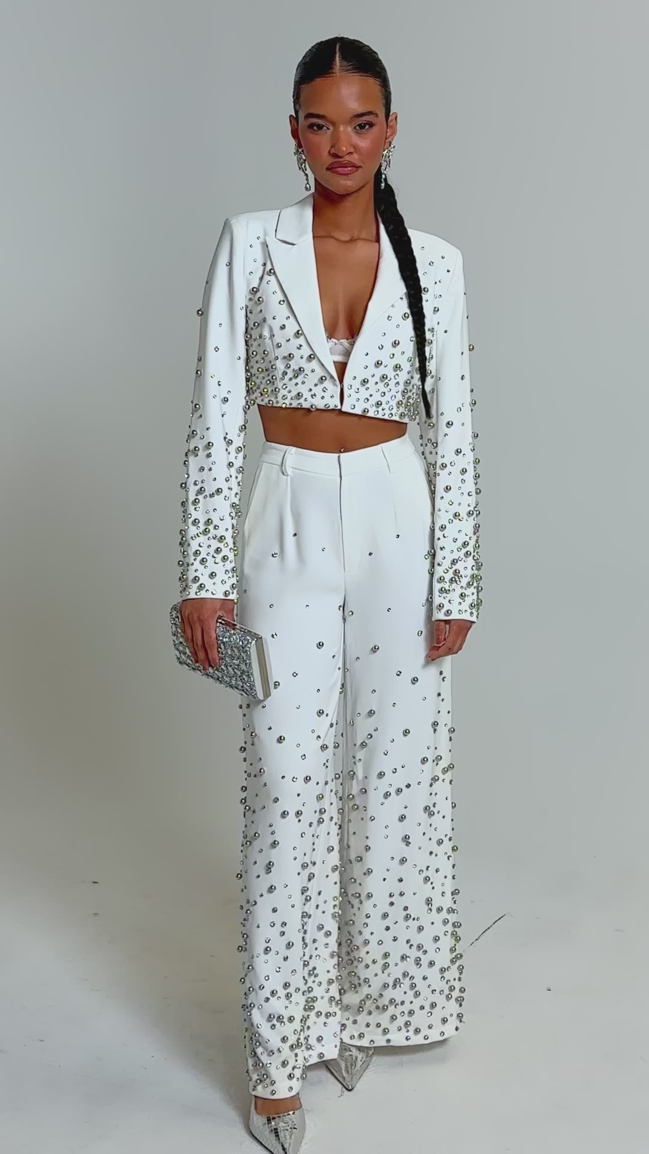 Video of woman modeling the GET CLOSER EMBELLISHED TROUSER IN WHITE, featuring pearl accents. She pairs the high-rise trousers with a cropped blazer, silver clutch, pointed heels, and wears her hair in a long braid for a chic look.
