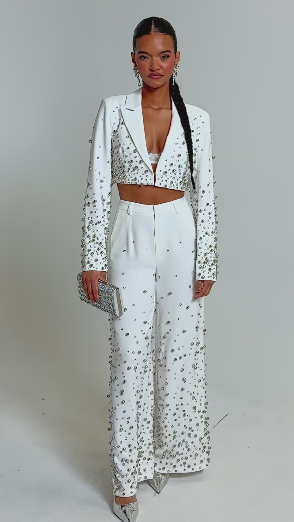 Video of woman modeling the GET CLOSER EMBELLISHED TROUSER IN WHITE, featuring pearl accents. She pairs the high-rise trousers with a cropped blazer, silver clutch, pointed heels, and wears her hair in a long braid for a chic look.