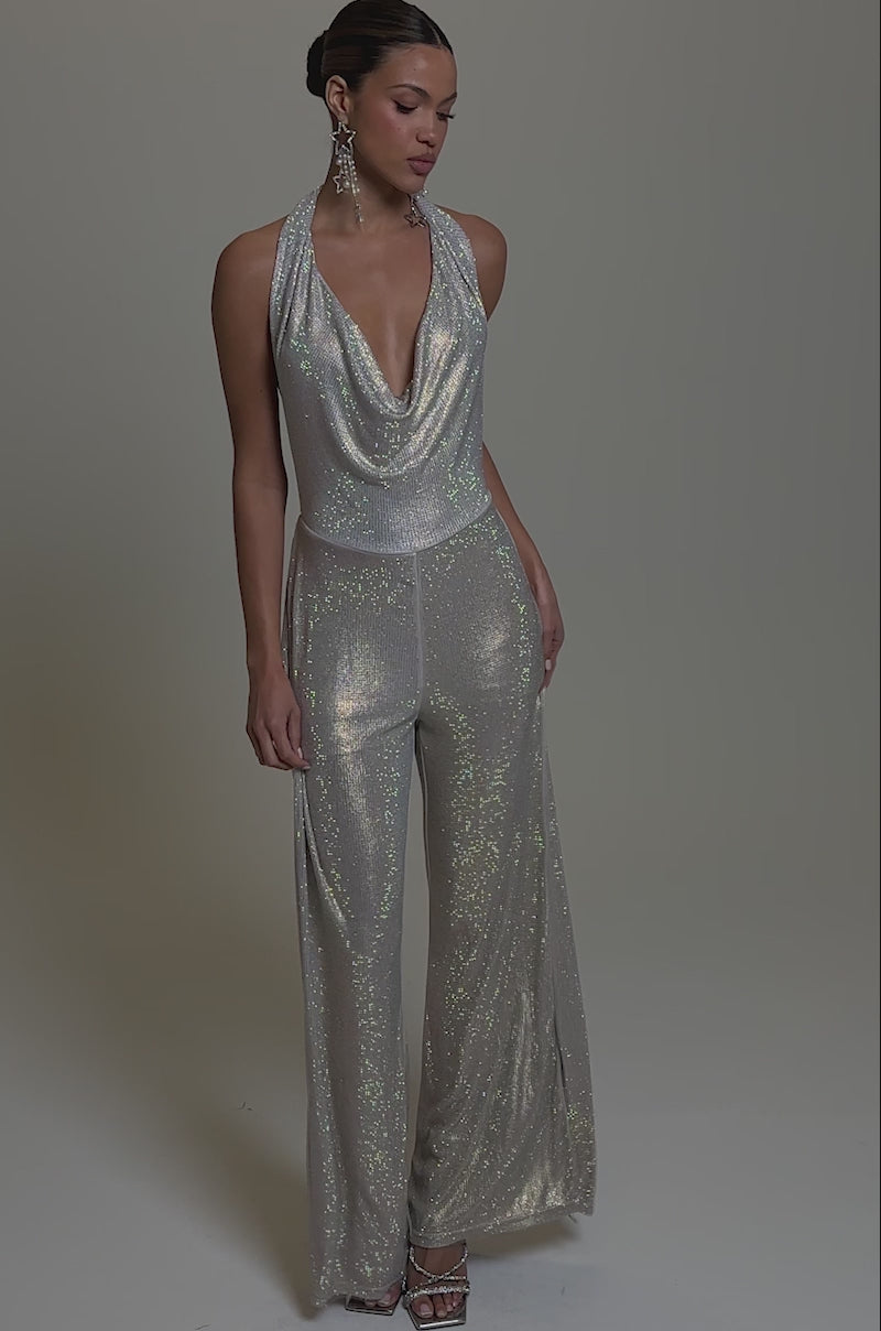 Video of woman modeling the MAKE YOU DO A DOUBLE TAKE RHINESTONE JUMPSUIT IN CHAMPAGNE, featuring a sleeveless, draped neckline and wide-leg pants. She wears slicked-back hair, statement earrings, and strappy heels against a plain gray background