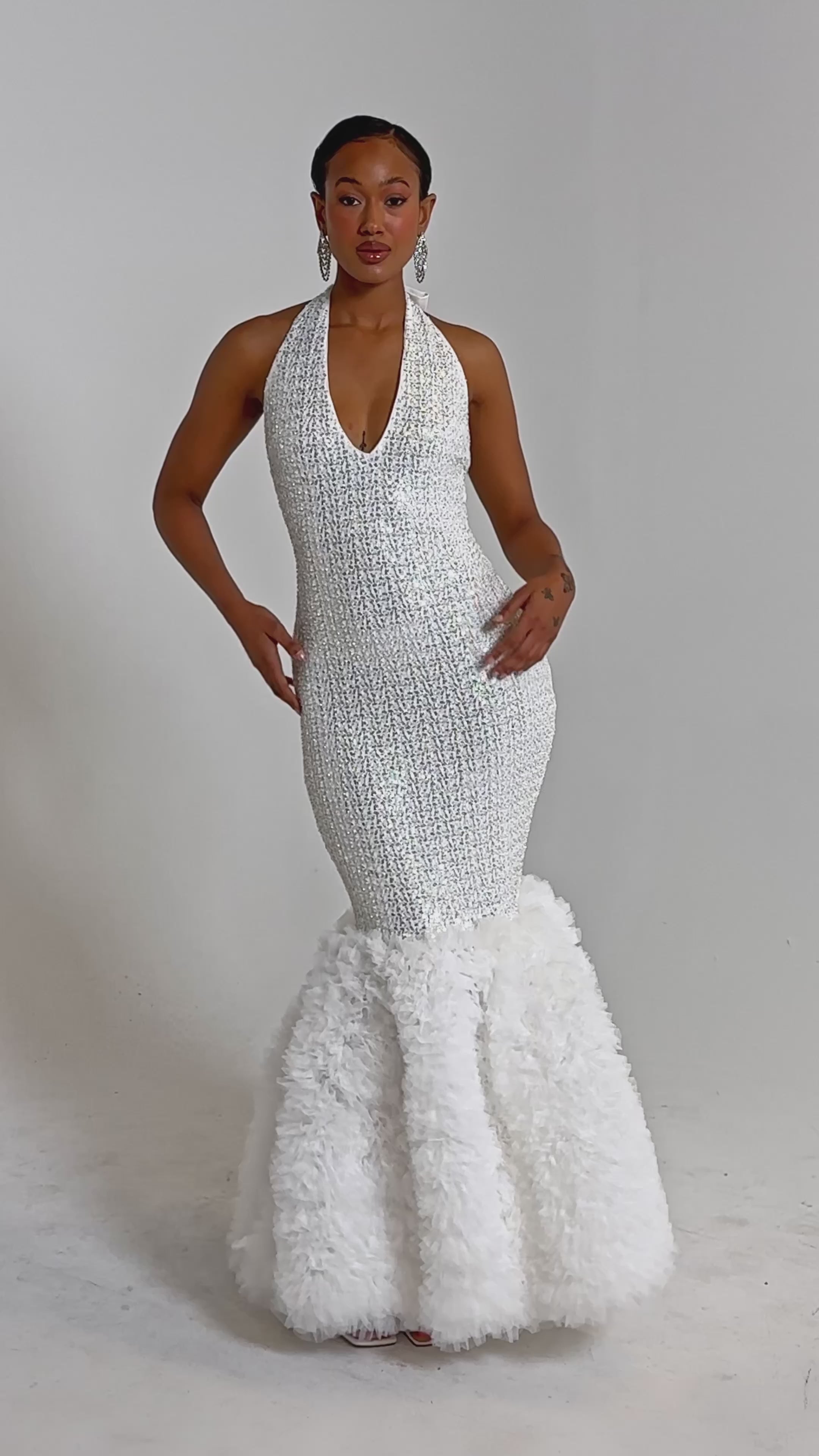 A video of a woman wearing the SHOWTIME SEQUIN MAXI DRESS, a sleeveless white mermaid gown featuring a halter neckline and a voluminous ruffled skirt.