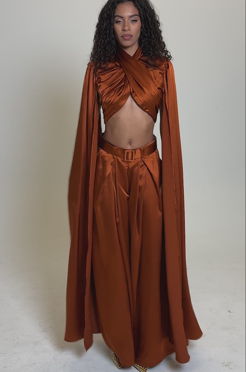 Video of woman modeling the ALL WE EVER KNEW TROUSER, wide-leg satin pants in burnt orange with a high-rise fit, paired with a matching draped crop top and cape. She accessorizes with gold shoes and jewelry, her curly hair styled naturally against a light background.
