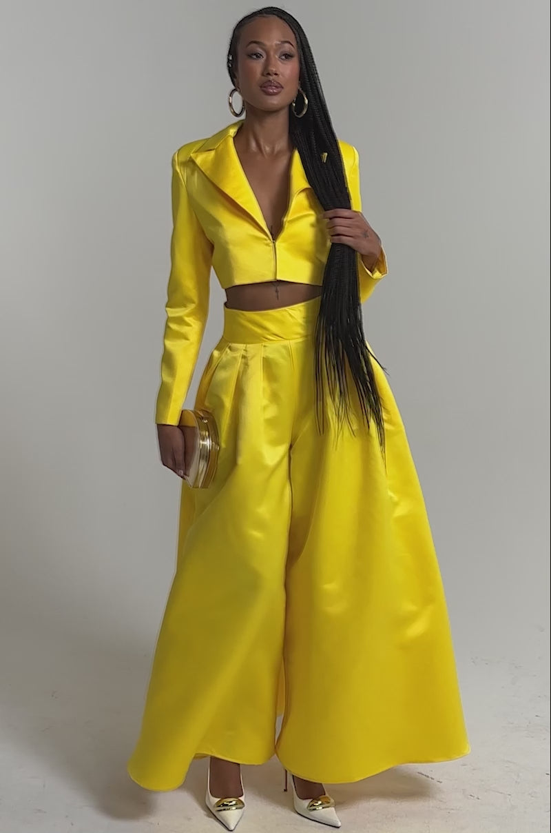 A video of a woman in the BLOOMING CROP SATIN BLAZER and matching wide-leg pants, holding a round gold clutch, wearing yellow heels.