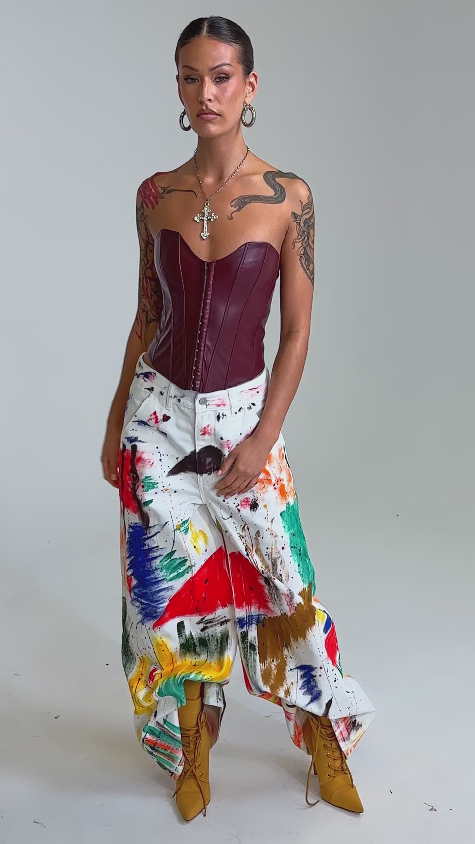 Video of woman modeling the PAINTED ON RELAXED DENIM PANT with colorful splatters, tan lace-up boots, large hoop earrings, and layered necklaces against a plain background.
