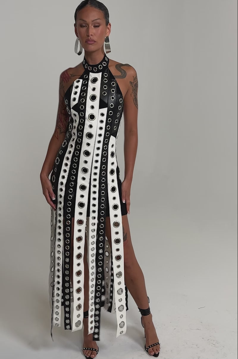 A video of a woman in the ON MY BEST BEHAVIOR TASSEL MAXI DRESS, featuring black-and-white vertical stripes, metal grommets, high slits, and open sections. 
