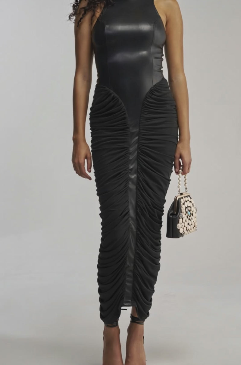 Video of woman modeling the YOU NEED ME RUCHED MAXI DRESS IN BLACK, which features a sleeveless design, fitted faux leather bodice, and bodycon silhouette. She holds a decorative handbag and wears black heels while standing against a plain backdrop.