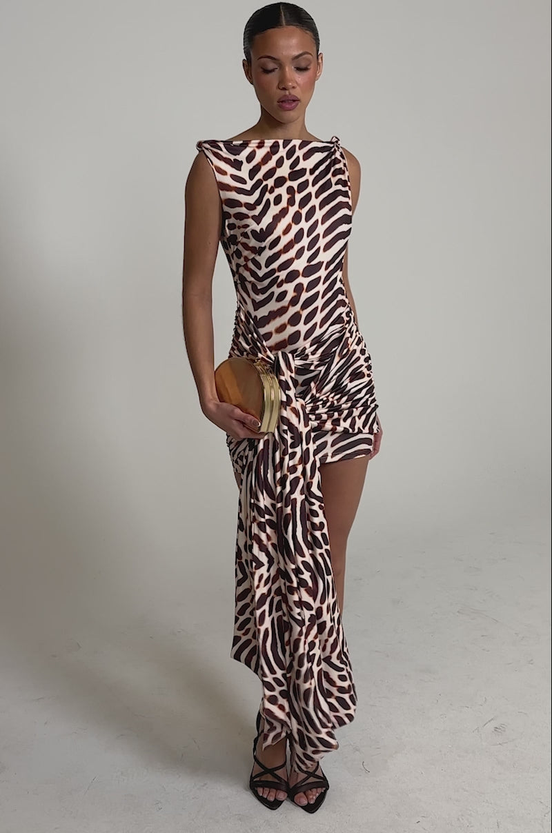 Video of woman modeling the JUNGLE TIES LEOPARD MINI DRESS—an off-the-shoulder, asymmetrical brown and beige animal-print mini dress—standing against a light gray background, holding a small clutch and wearing brown strappy heels with her hair in a long braid.