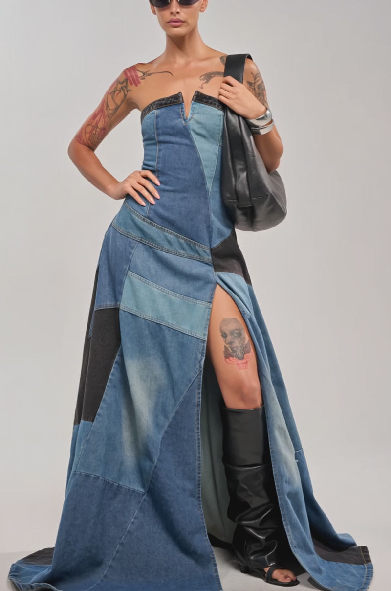 Video of woman modeling the LIFE IN DENIM STRAPLESS MAXI DRESS, featuring patchwork details and a high slit. Styled with black heeled boots, sunglasses, and sleek hair, this look is completed by a black leather accessory over one shoulder.