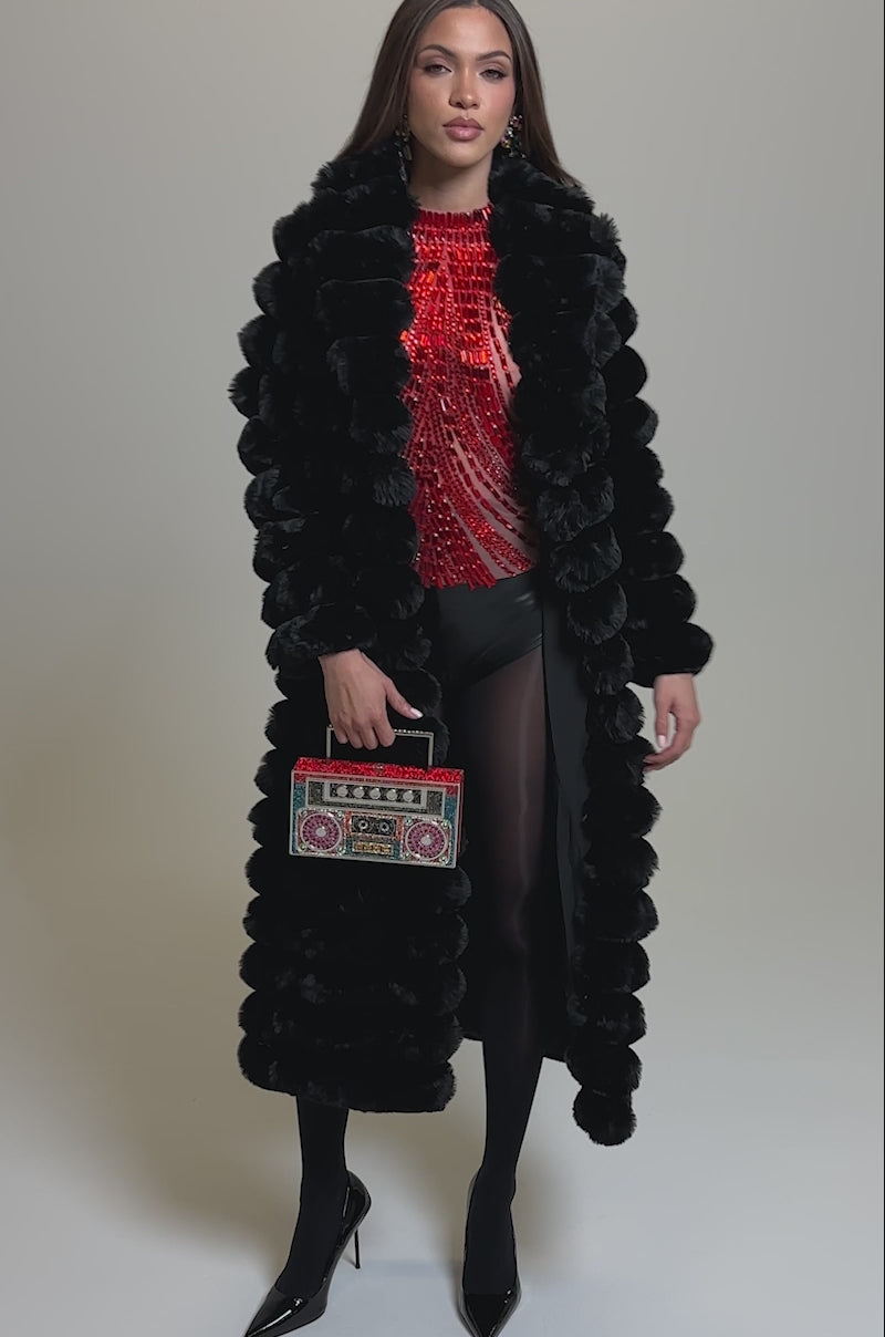 Video of woman modeling the MAXI POPCORN FAUX FUR IN BLACK over a red top, holding a vibrant box-shaped handbag. She has straight brown hair, wears black heels, and is posed against a plain light background.