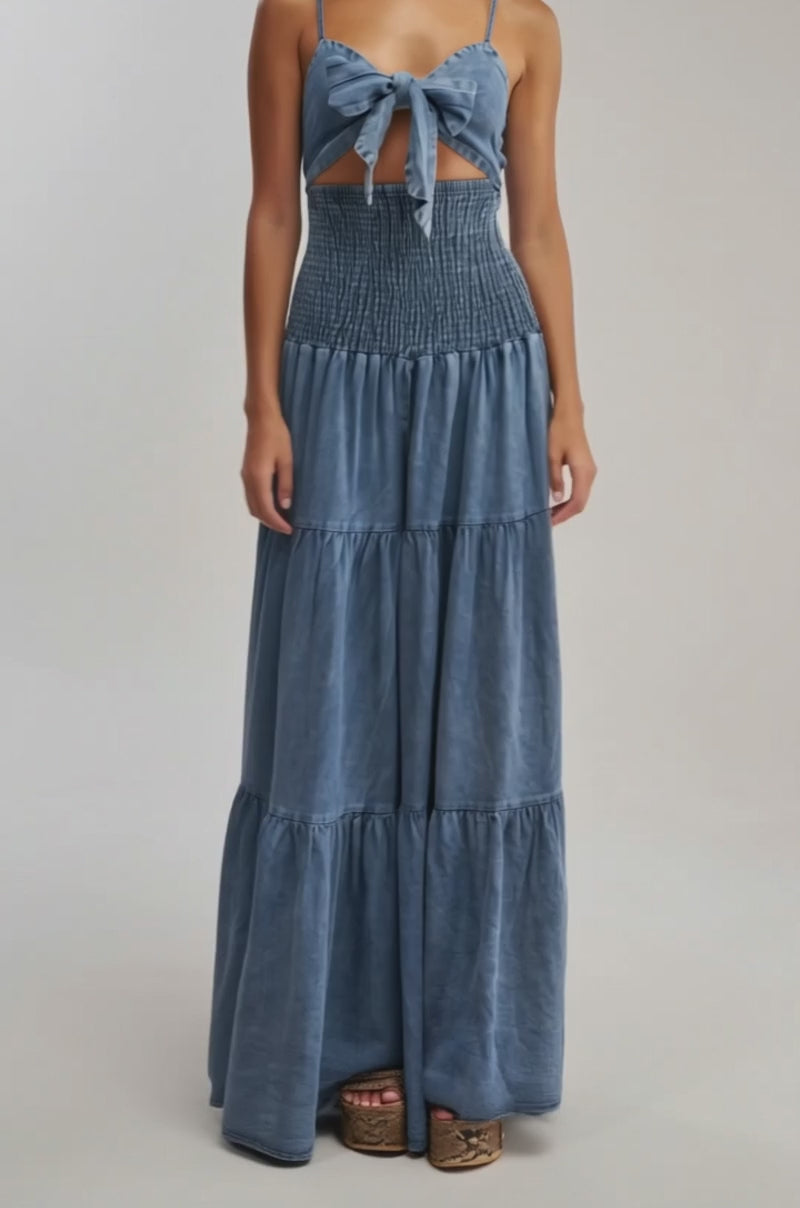 Video of woman modeling the JUMPIN AROUND DENIM JUMPSUIT, a sleeveless denim outfit with thin straps, front tie detail, smocked waist, and tiered legs—perfect for vacation—against a plain light background.