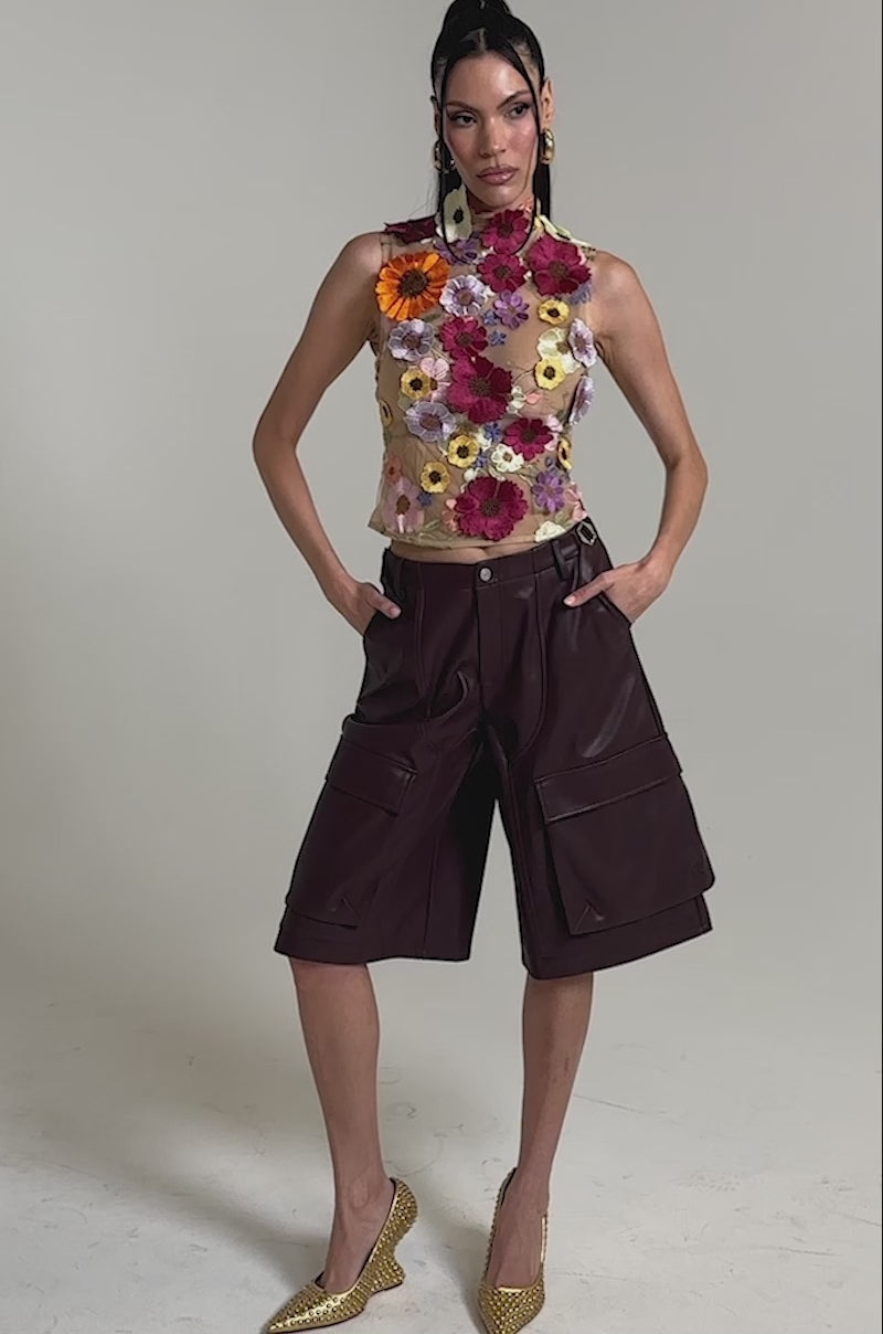 A video of a woman in the WHERE THIS FLOWER BLOOMS MESH TOP, featuring a sheer mesh design with colorful 3D floral appliqués, styled with burgundy leather knee-length shorts, gold studded heels, and gold accessories against a plain background.