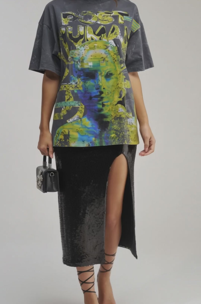A video of a woman wearing the BETTER DAYS TO COME OVERSIZED GRAPHIC TEE with a rhinestone abstract yellow and green print, paired with a black sequin high-slit skirt, strappy black heels.